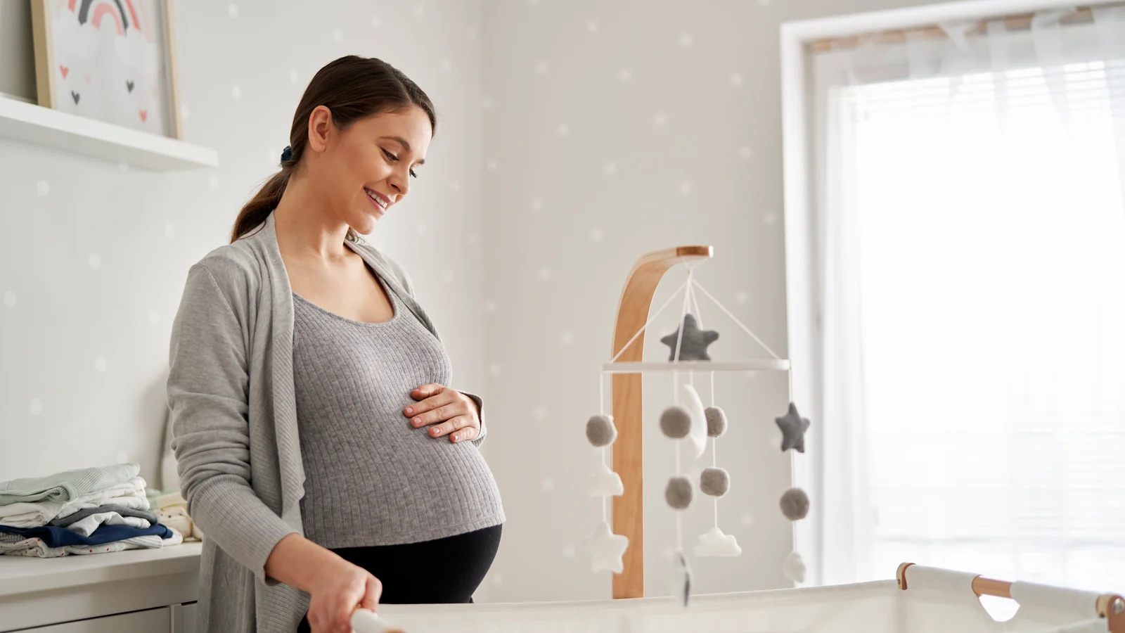 Smiling pregnant woman with a hand on her belly beside a white bassinet in a bright, simple Montessori nursery