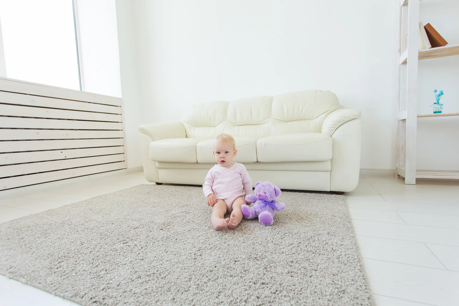 Baby in a pink bodysuit sitting on a soft rug beside a purple teddy bear in a bright room with plain white walls