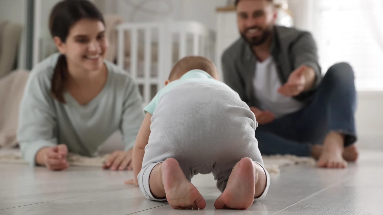 Two smiling parents on a bright wood floor watch a baby crawl toward them: a Montessori observation journal starts with watching