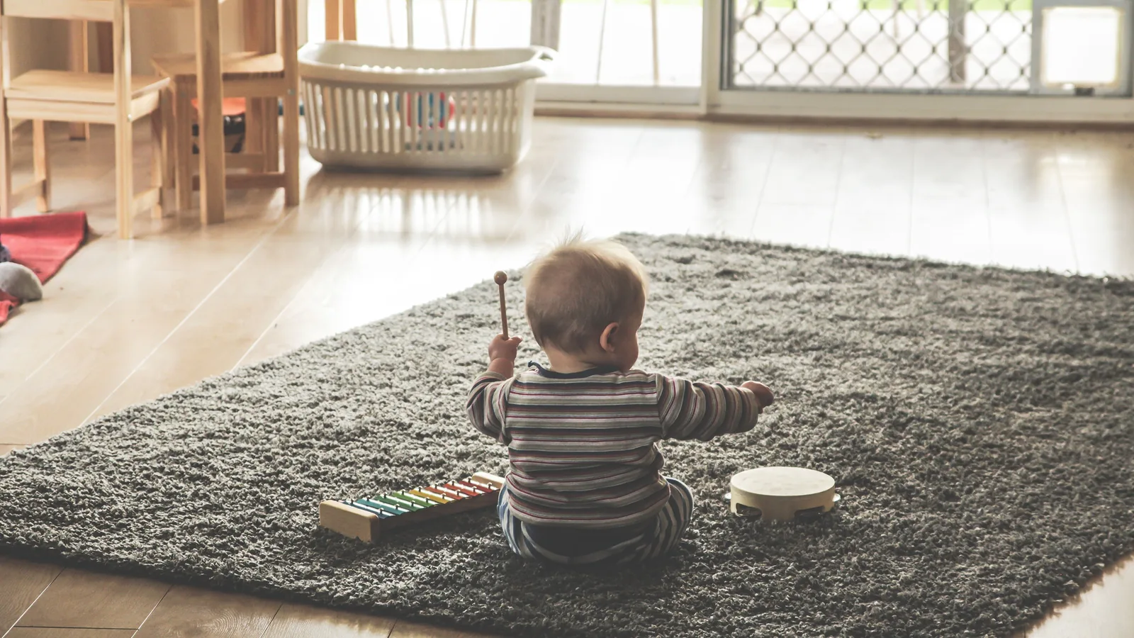 A baby in a striped top plays with a toy xylophone and a tambourine on a living room rug: Montessori on a budget