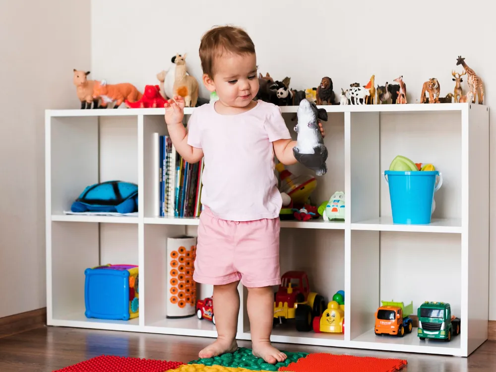 A toddler stands by a low cube shelf with books, toy animals and baskets within reach