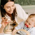 Mother holding a wooden stacking ring while a baby on a bed watches the toy: a Montessori presentation