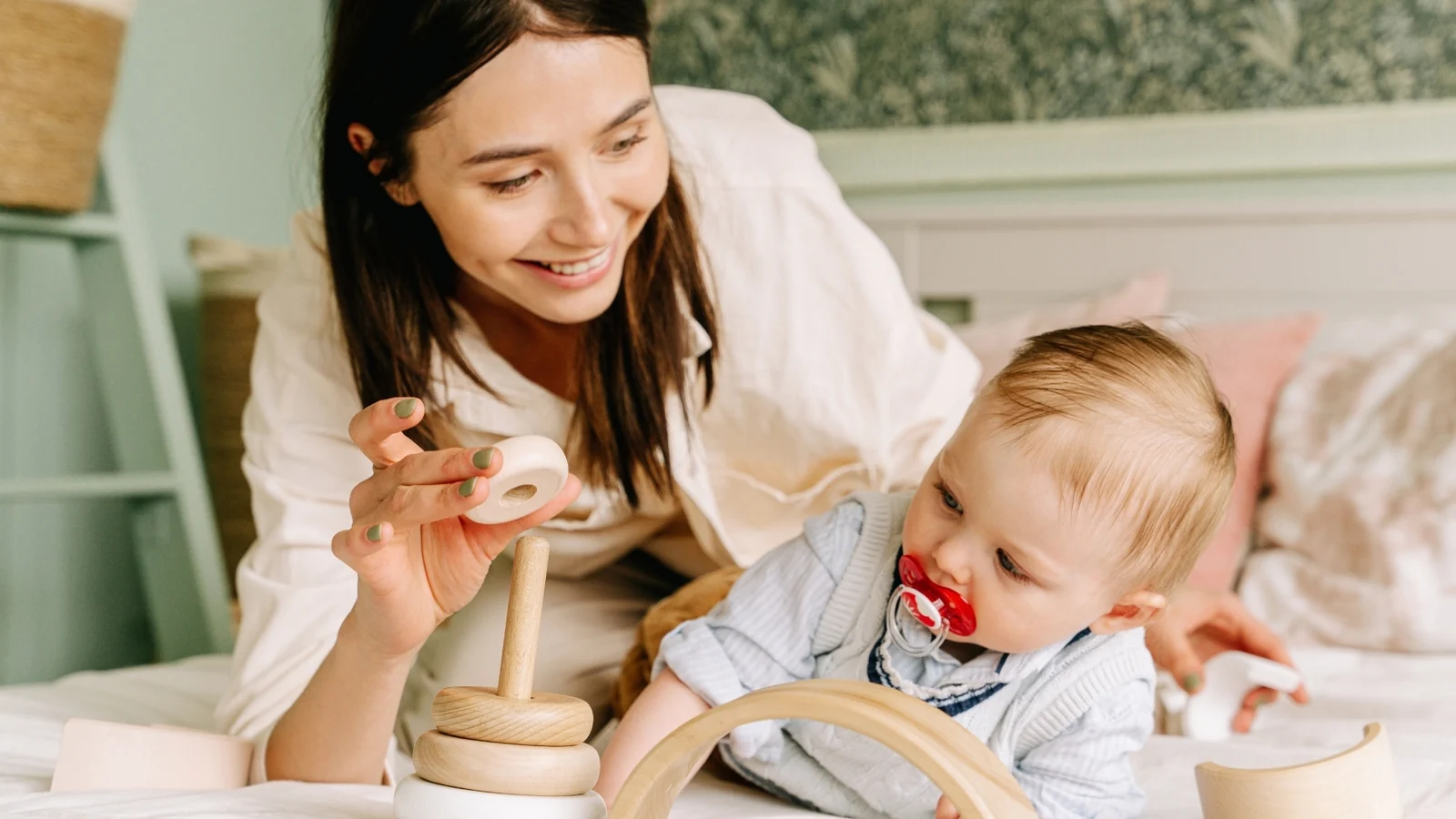 Mother holding a wooden stacking ring while a baby on a bed watches the toy: a Montessori presentation
