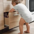 A toddler in a gray bodysuit stands at an open wooden kitchen drawer and reaches inside