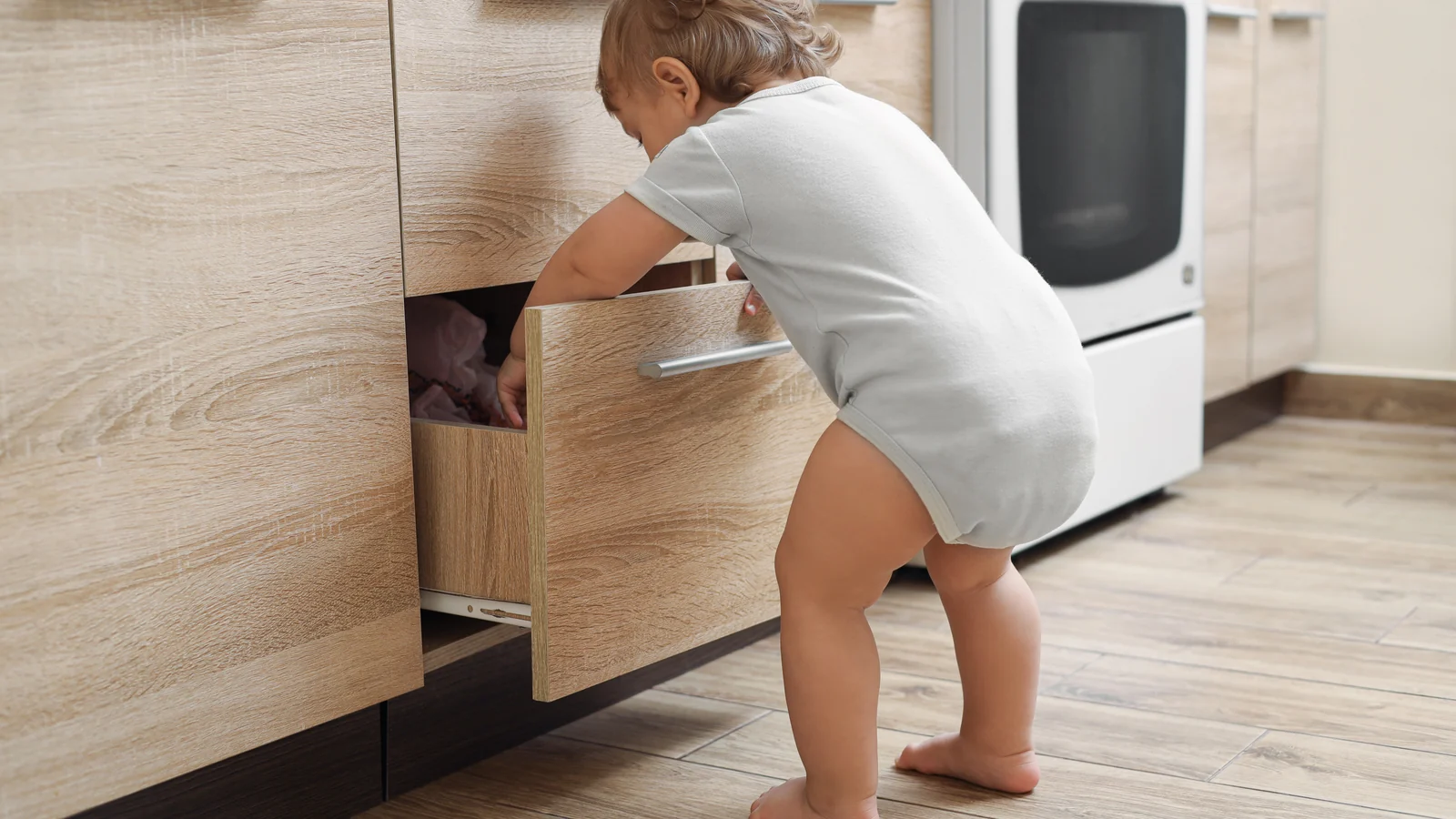 A toddler in a gray bodysuit stands at an open wooden kitchen drawer and reaches inside