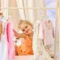 Smiling toddler in an orange dress sitting by a wooden clothes rack, one hand up to a hanger, a Montessori wardrobe corner