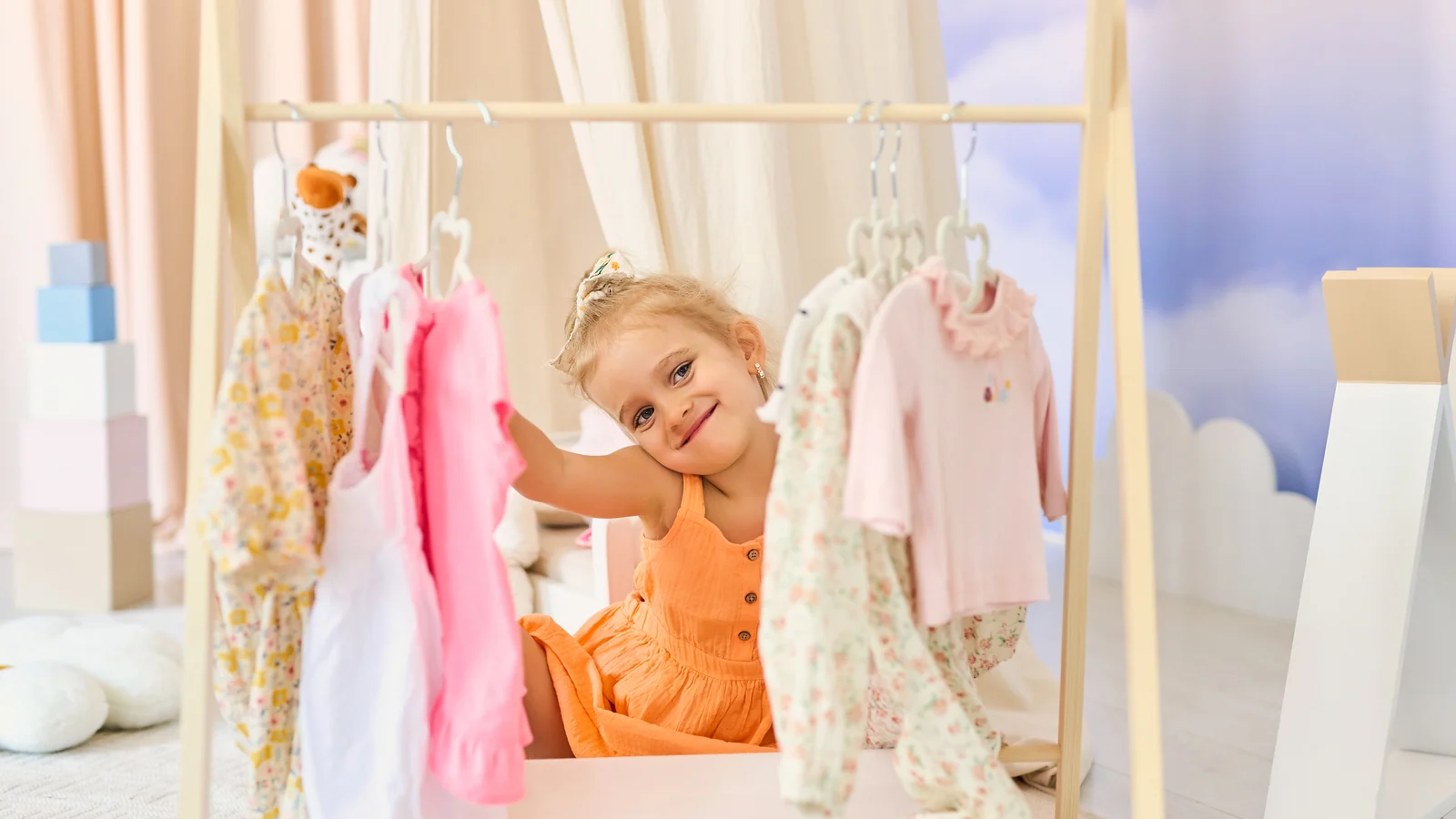 Smiling toddler in an orange dress sitting by a wooden clothes rack, one hand up to a hanger, a Montessori wardrobe corner