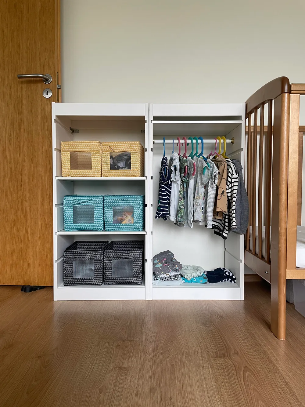 Short white open wardrobe with a hanging rail of kids' clothes, fabric storage boxes and folded clothes beside a wooden crib