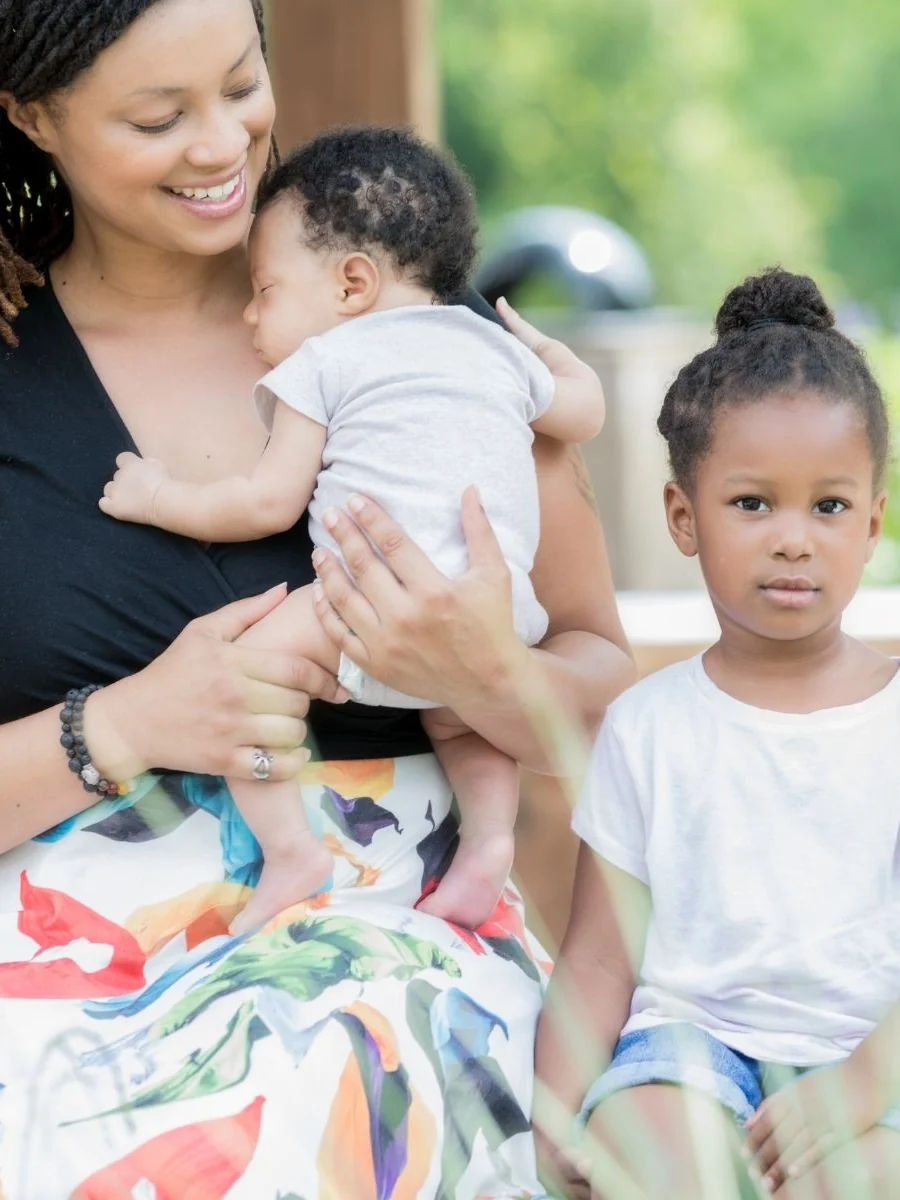 Smiling mother holding a newborn against her chest while a preschool-age girl sits close beside her outdoors