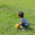 Toddler in a striped shirt crouching in the grass, looking down at a yellow ball: nature scavenger hunt for toddlers
