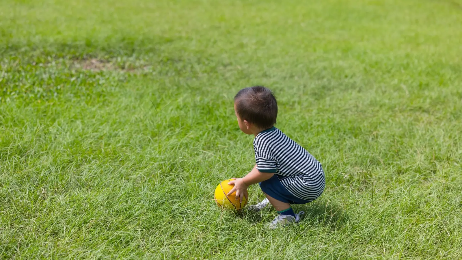 Toddler in a striped shirt crouching in the grass, looking down at a yellow ball: nature scavenger hunt for toddlers