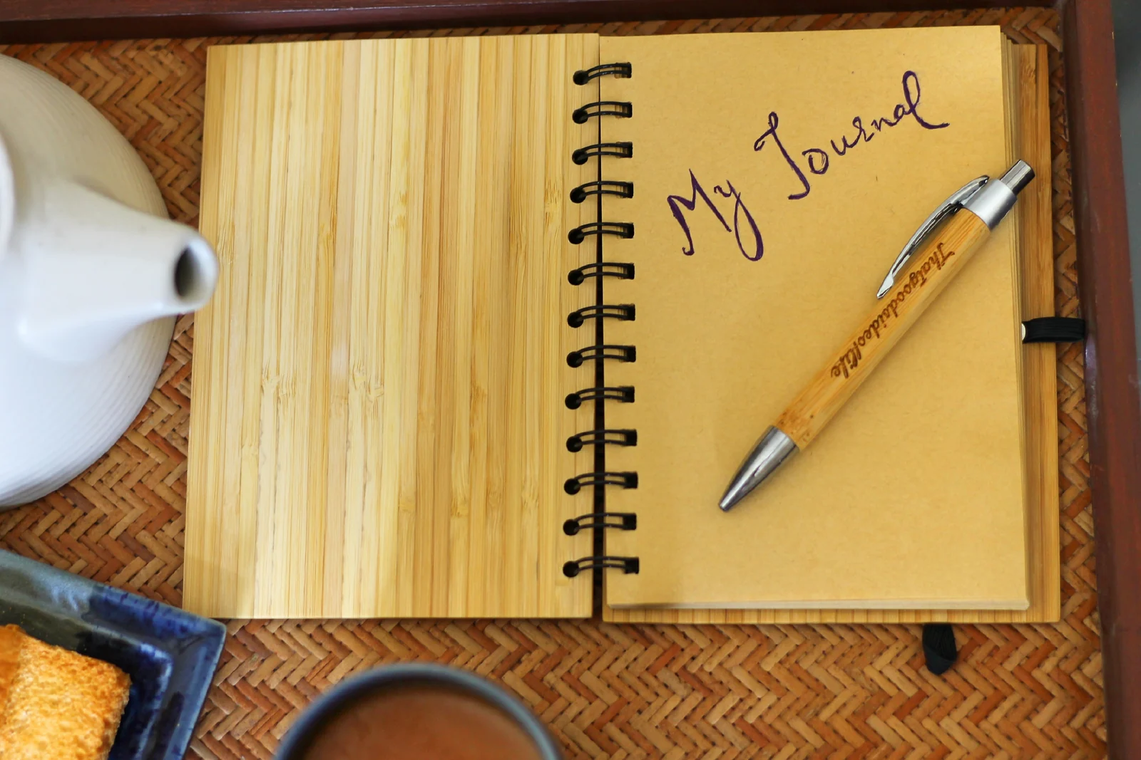 Open journal with a pen beside a teapot on a woven tray