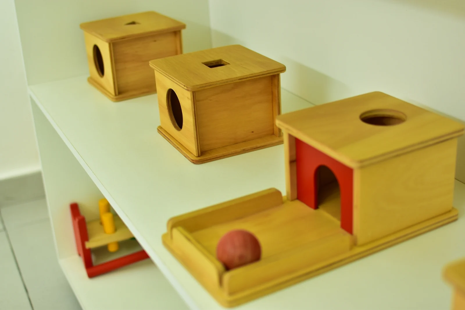 Wooden object permanence boxes on a low white shelf, with a red wooden ball in the tray of the largest box
