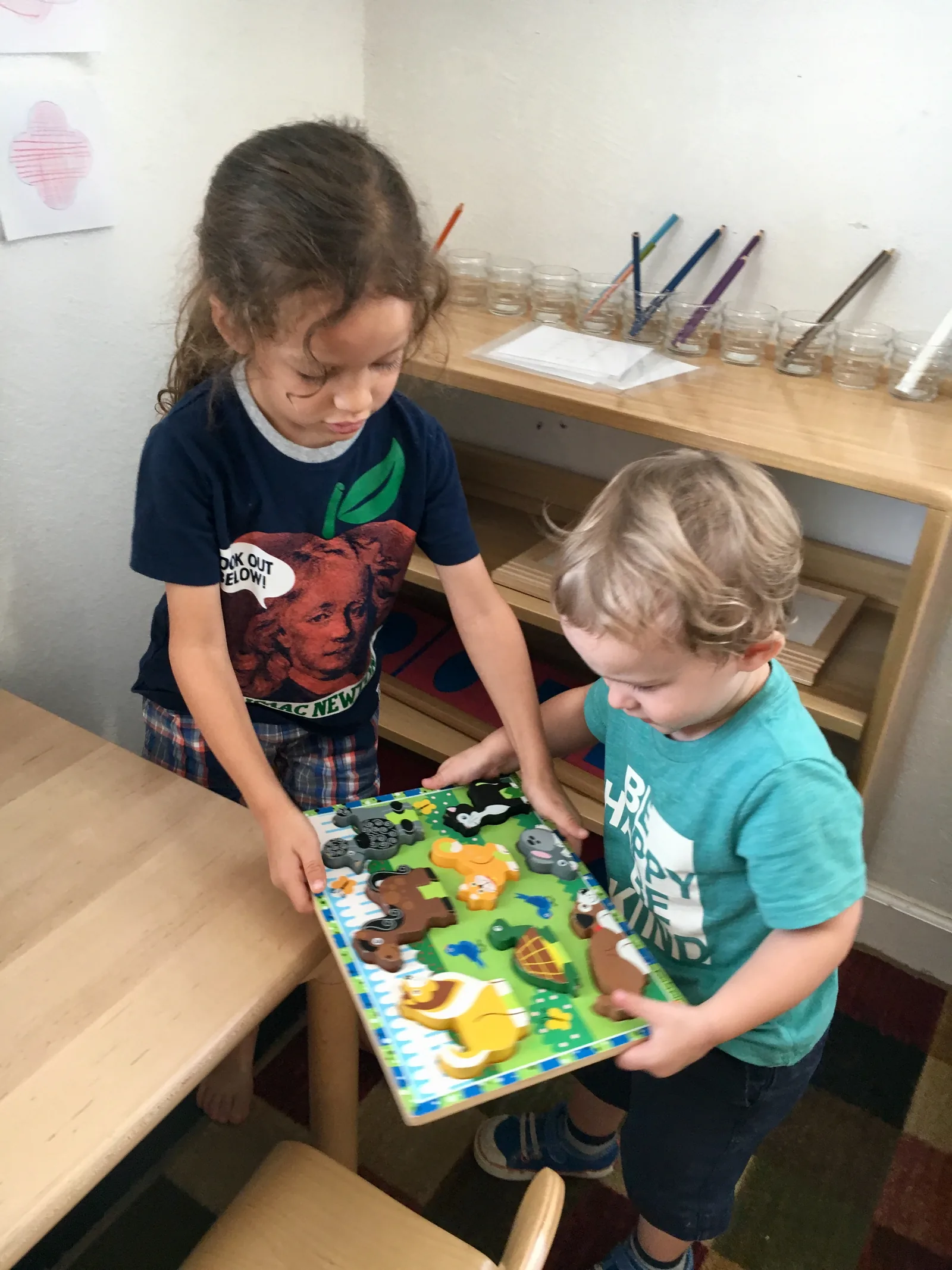 Older girl and a younger boy carrying a wooden animal puzzle together beside a low shelf in a classroom