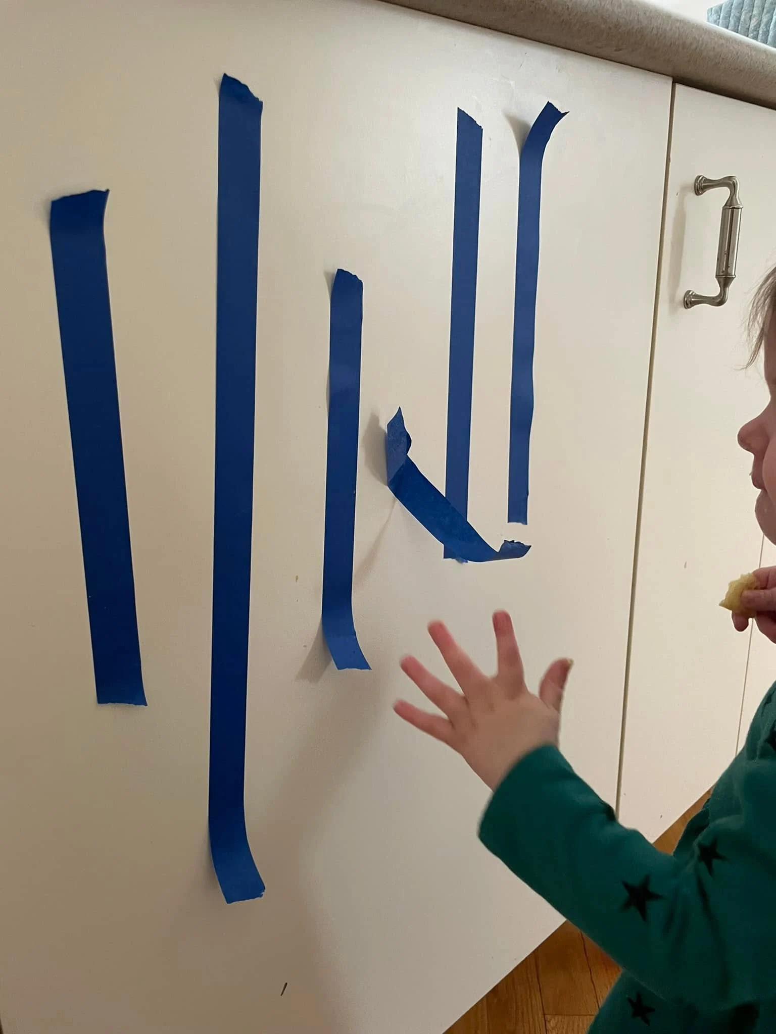 Strips of blue painter's tape on a white kitchen cabinet door, with a toddler's hand reaching toward them