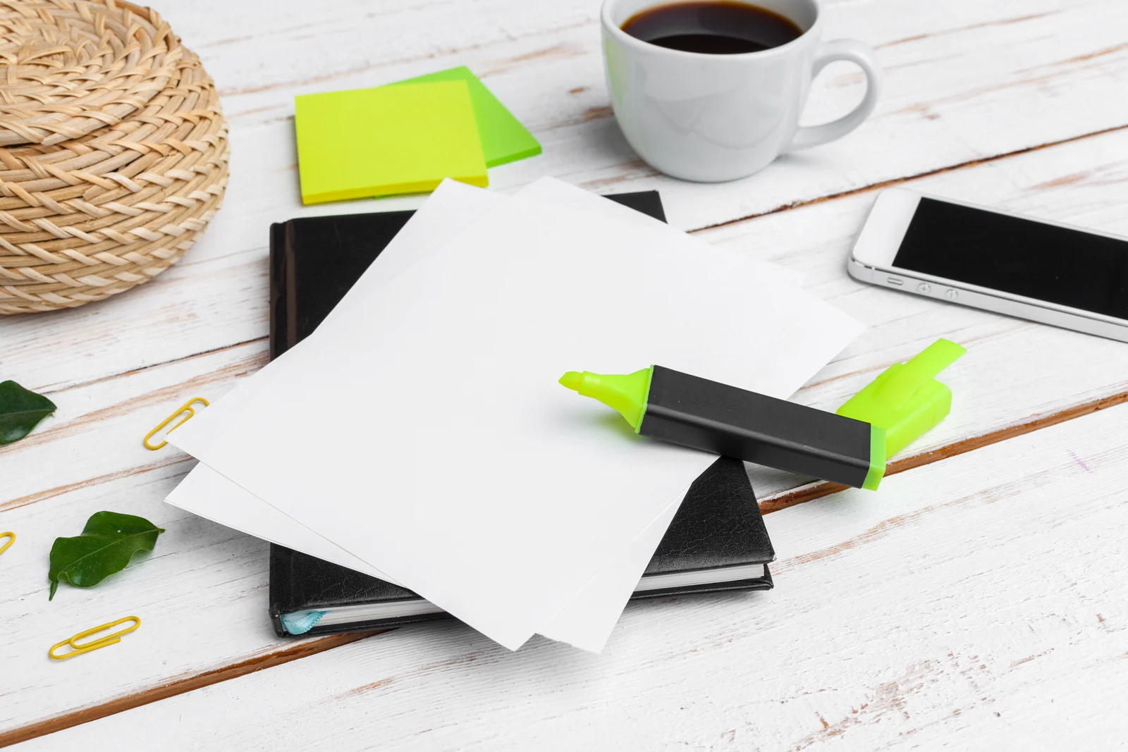 Loose sheets of paper and a highlighter on a notebook, on a white wooden table beside a cup of coffee and a phone