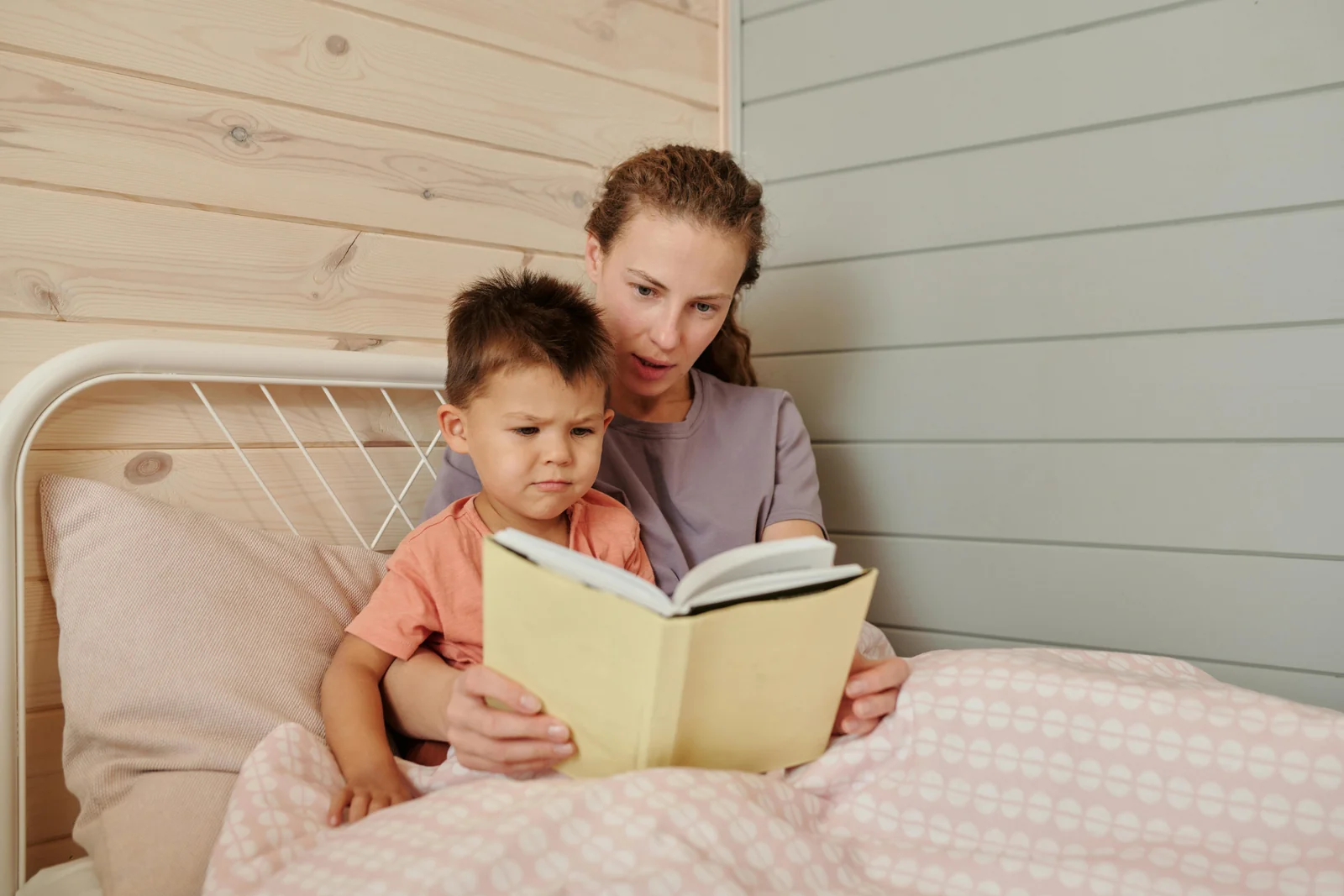 Parent and toddler sitting up together in bed under a pink patterned blanket, reading a book