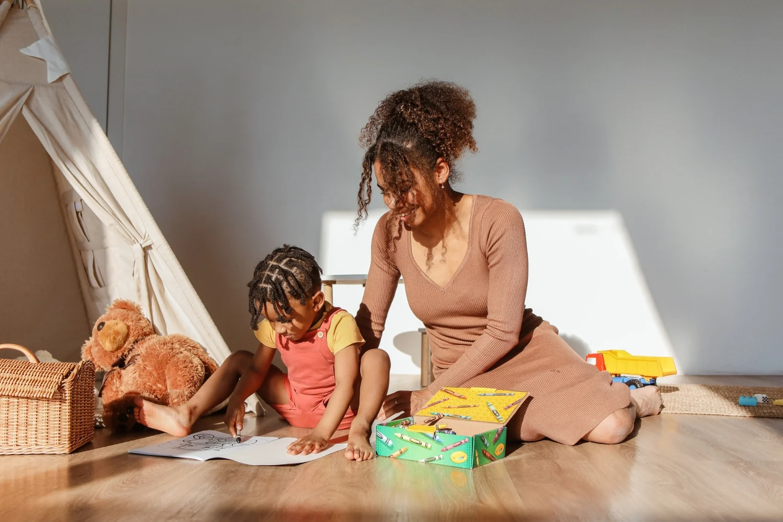 A parent and a toddler sit together on a wooden floor beside a play tent, looking at a box and a book