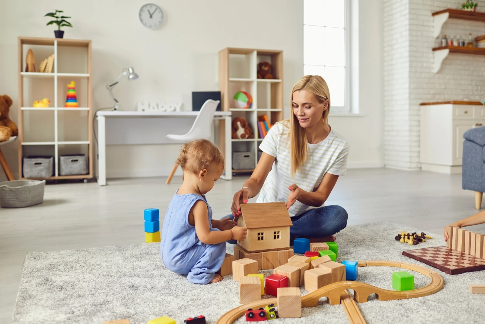 Parent and toddler building with wooden blocks next to a toy train track on a rug in a living room