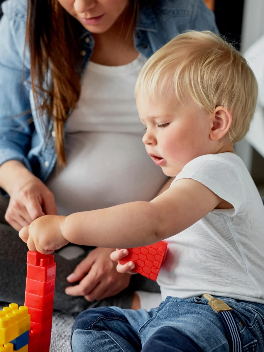 A pregnant woman and a toddler building a tower of big plastic blocks together on the floor