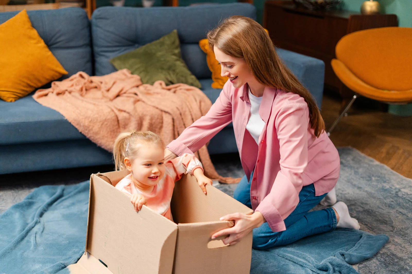 A mother kneels on a rug beside a baby sitting inside a cardboard box in a living room