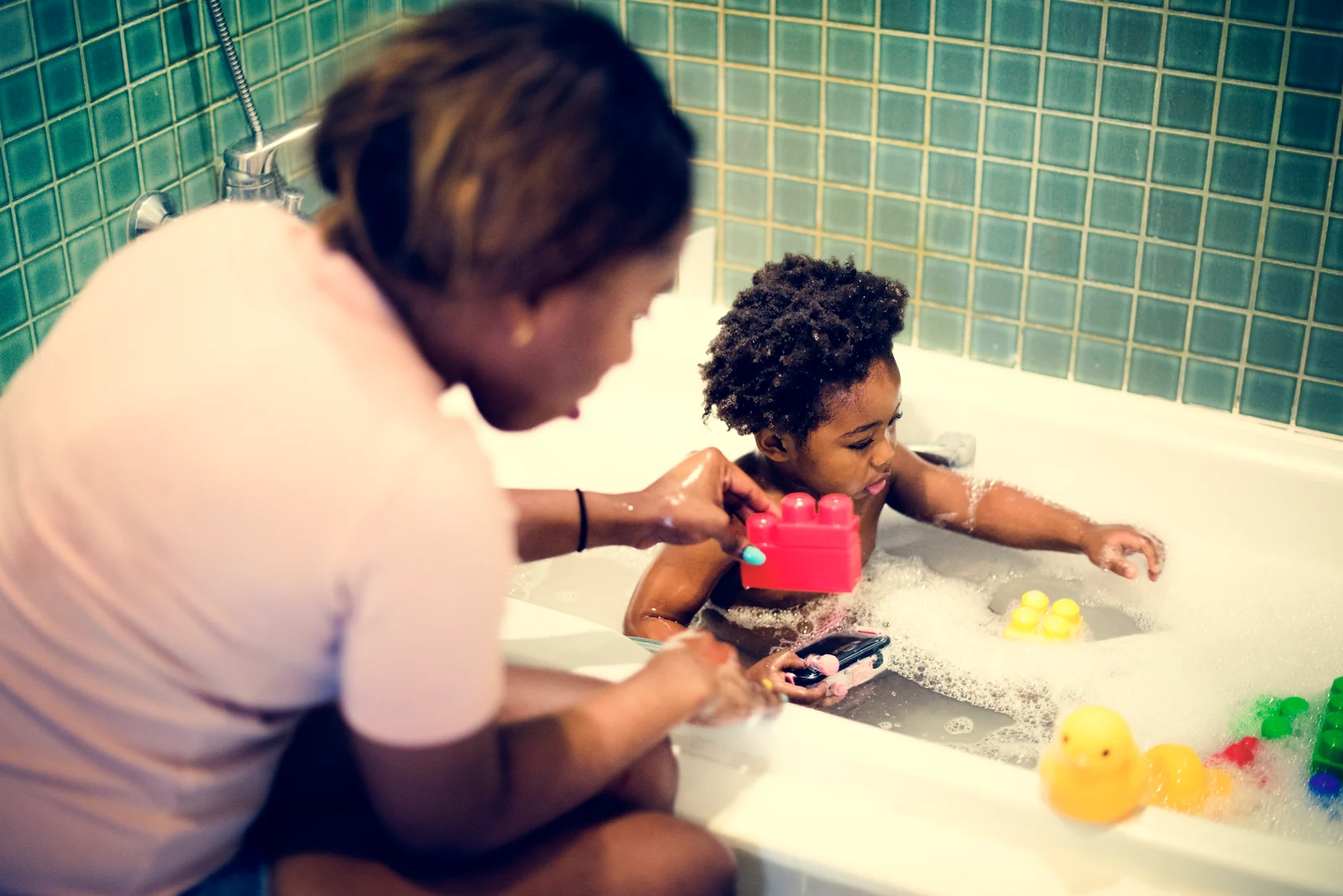 Parent kneeling beside a bathtub, holding a pink toy brick toward a toddler playing in the bubbles with rubber ducks