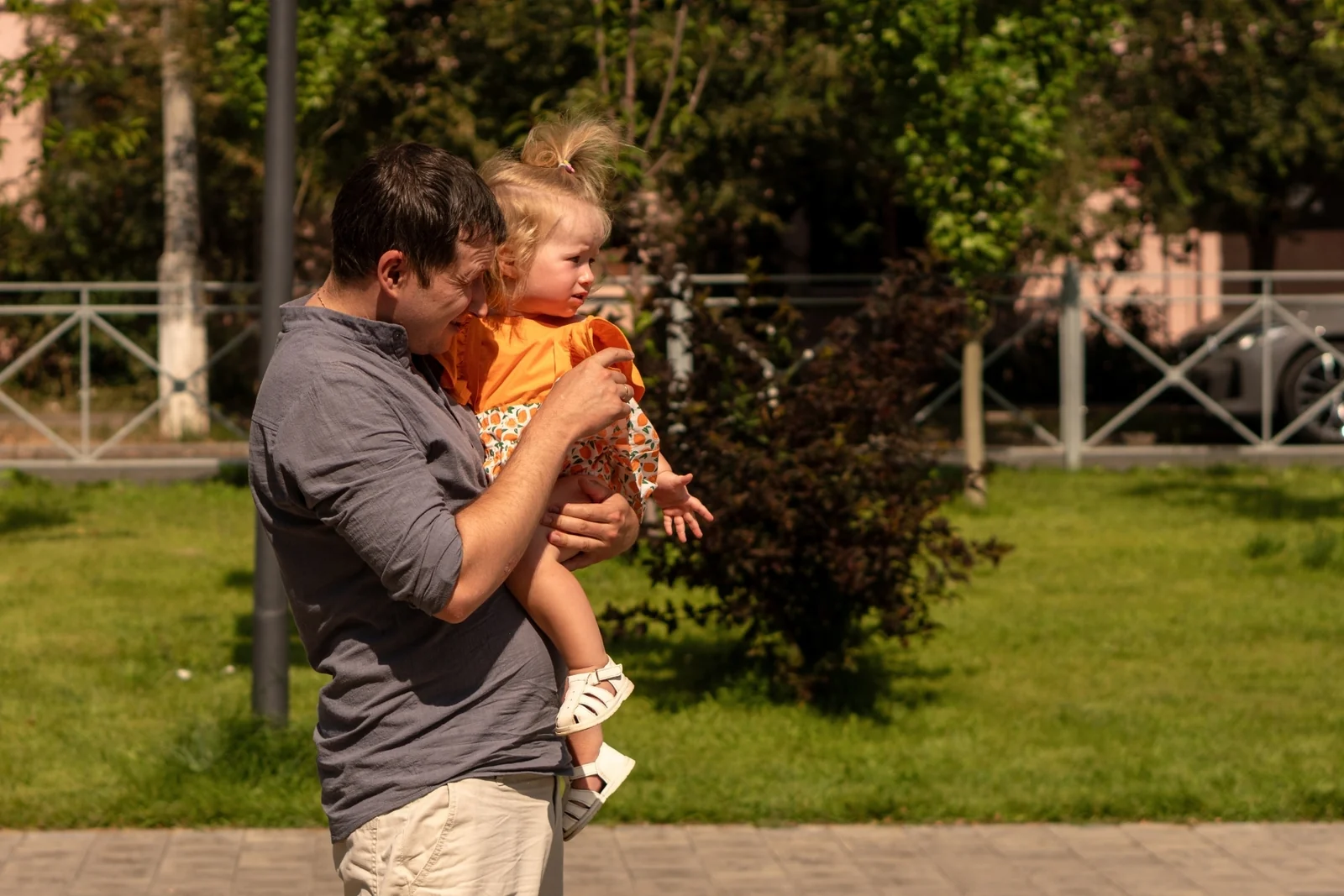 Man carrying a toddler on one arm and pointing ahead as they walk past a grassy lawn in a park