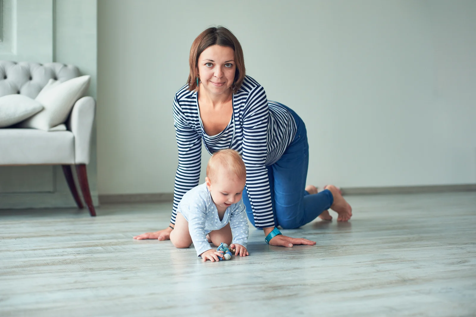 A parent on hands and knees on a plank floor, crawling beside a baby who reaches for a crocheted toy