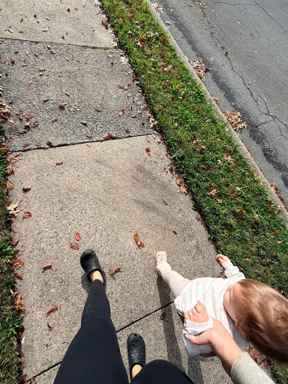 Parent in black clogs on a sidewalk with one hand under a baby in a white sweatshirt, with a grass strip and road beside them