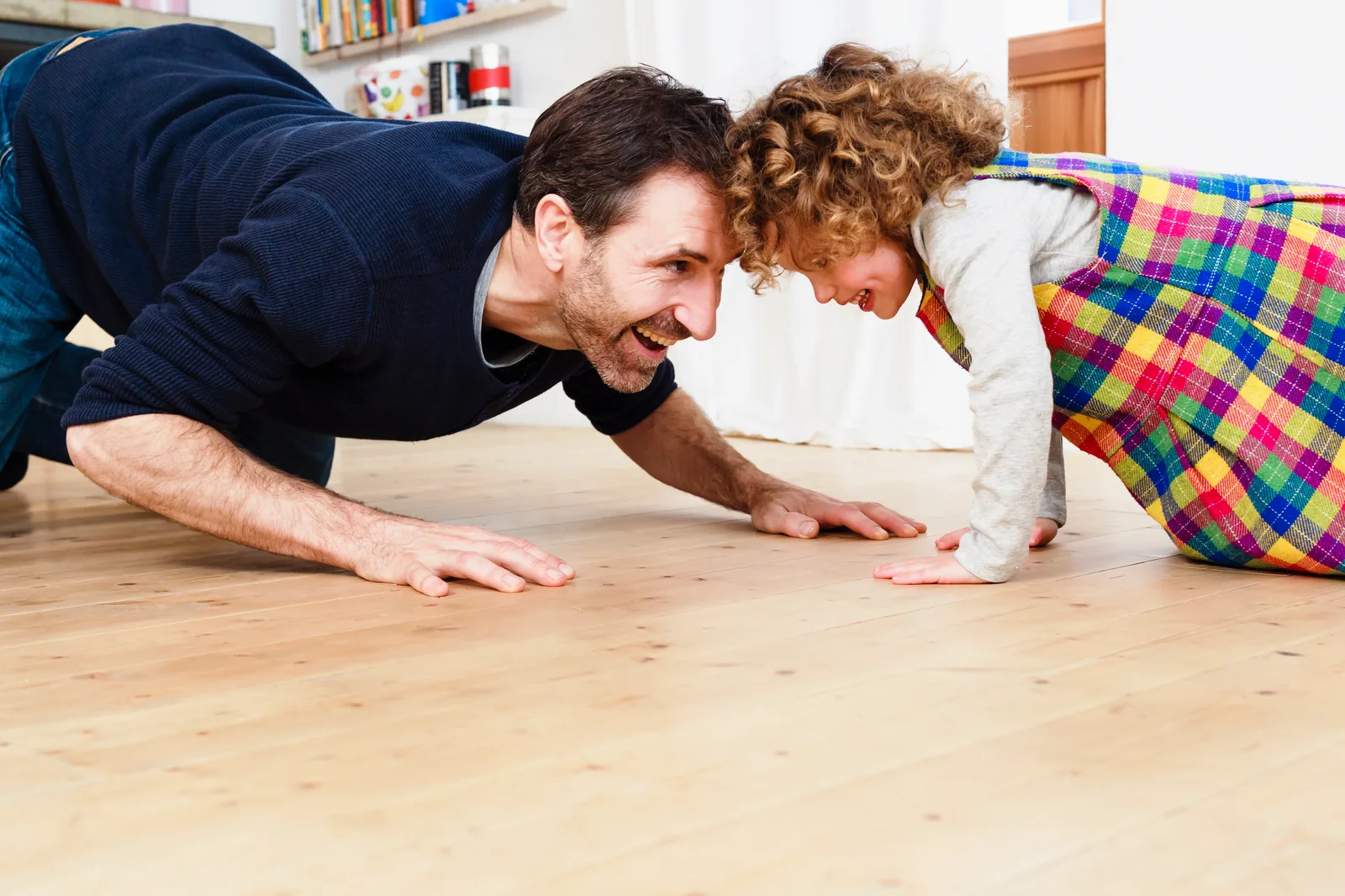 A parent on hands and knees on a wood floor, face to face with a smiling toddler at the child's eye level