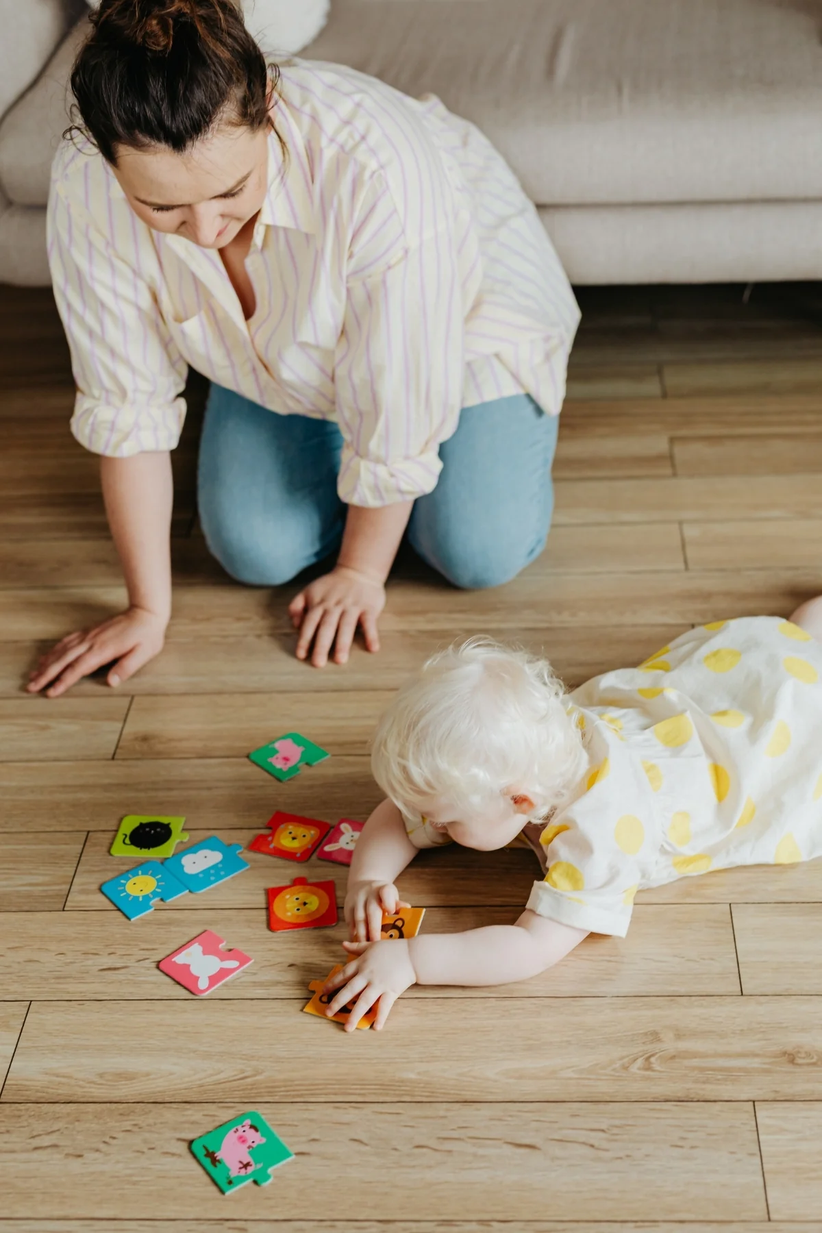 Woman kneeling on a wood floor with her hands down, watching a baby reach for colorful animal puzzle pieces