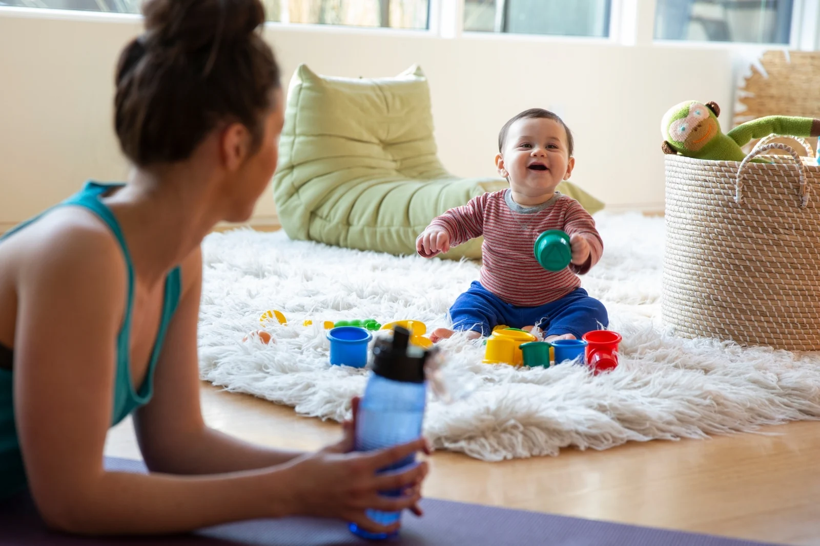 A parent sits on a couch with a mug, watching a baby play on a rug below