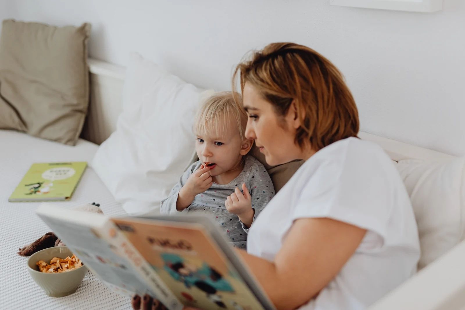 A parent reading a picture book to a toddler who is snacking, both sitting on a white bed in soft daylight