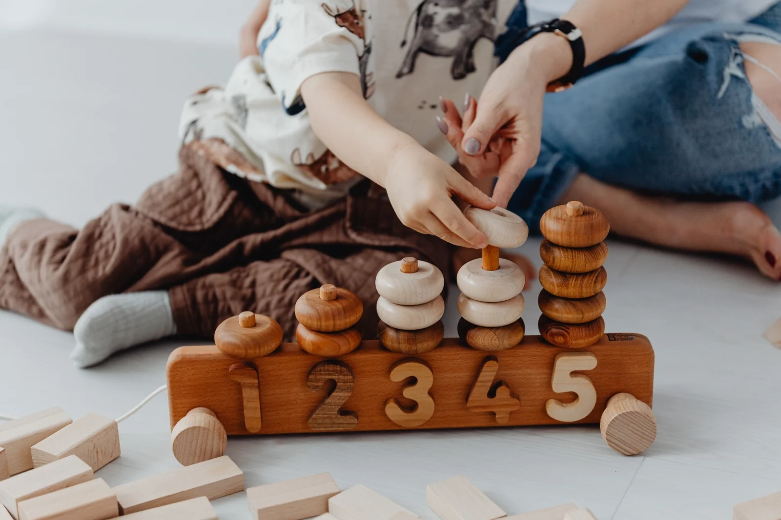Close-up of a parent's hand slowly placing a wooden ring on a counting toy while a toddler sits beside them