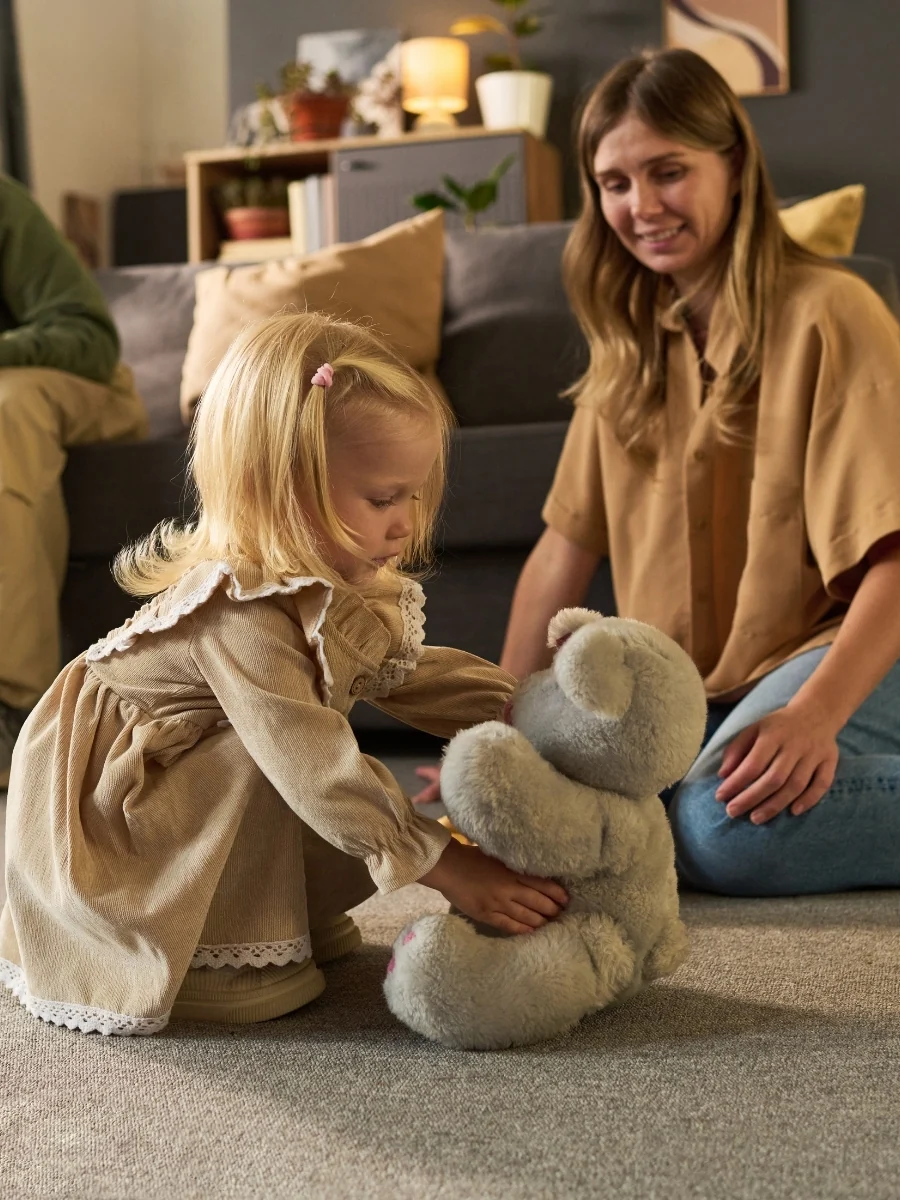 A parent sits on the floor a short distance away while a toddler plays on a rug