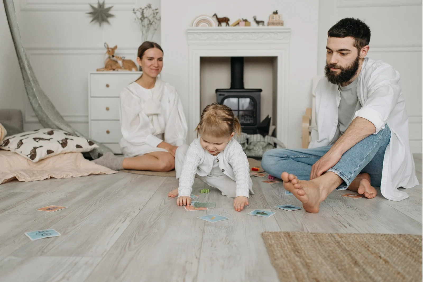 Parent sitting on the floor a little way back, watching a toddler play with a wooden toy