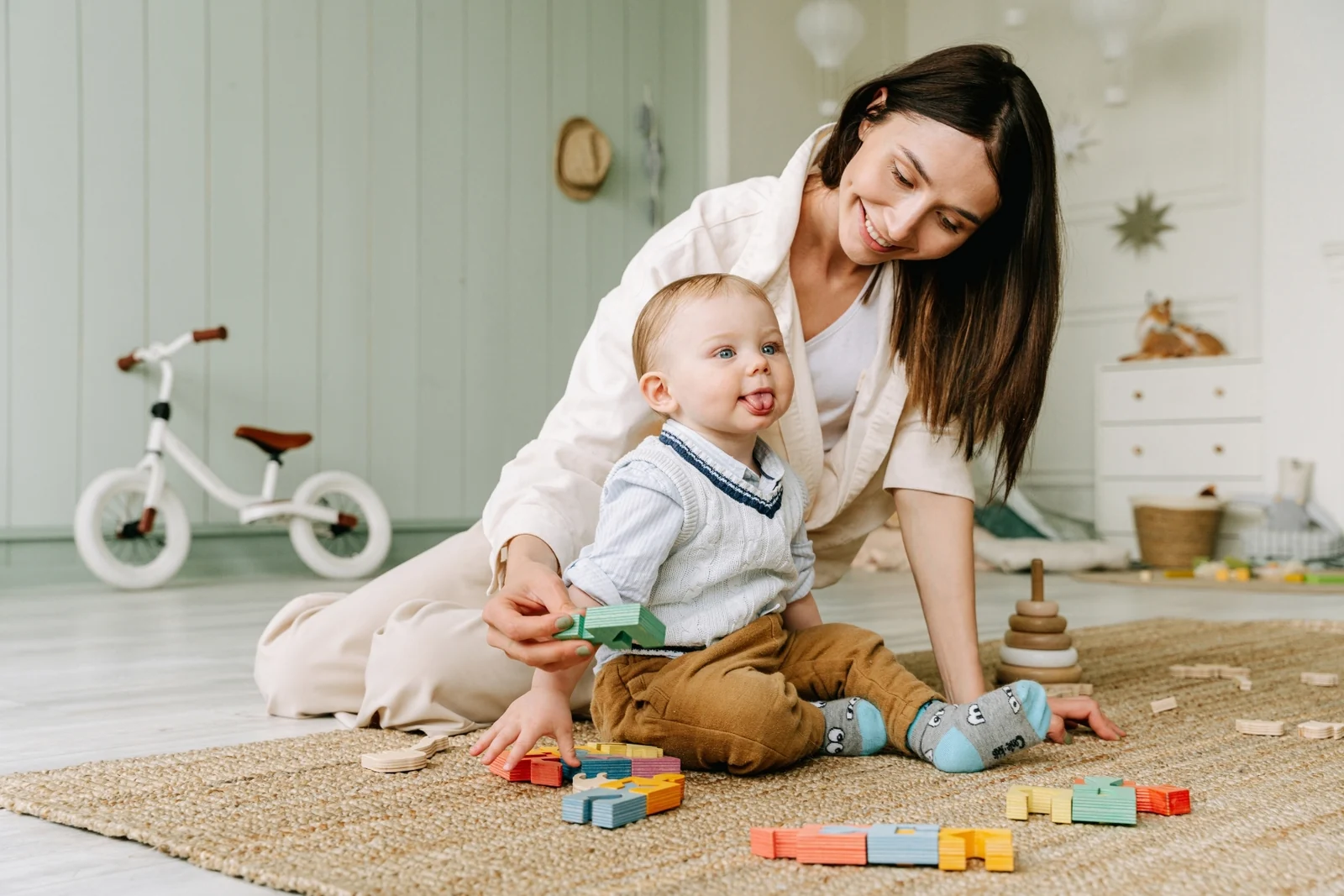 A smiling parent kneels on a woven rug beside a baby who sits and plays with colorful wooden blocks