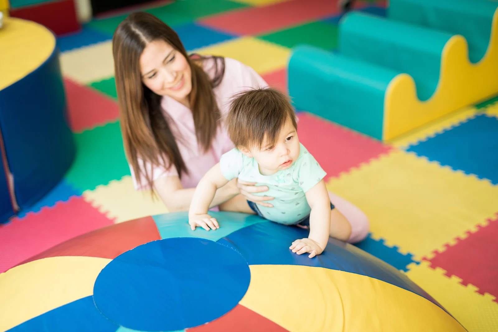 Parent sitting back on the floor, quietly watching a baby explore a large wooden toy on a play mat