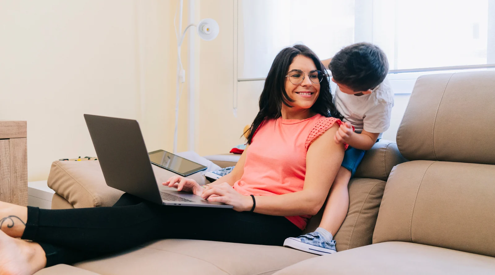 Woman working on a laptop on the sofa, smiling at a young child leaning on her shoulder
