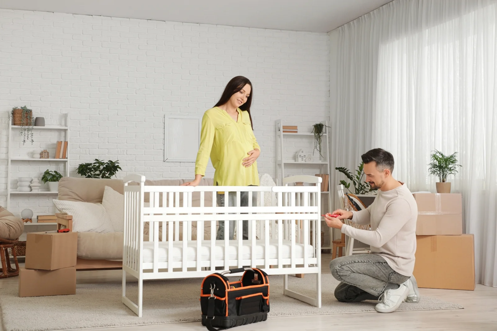 Expecting couple putting together a white crib in a bright room, a toolbag on the floor