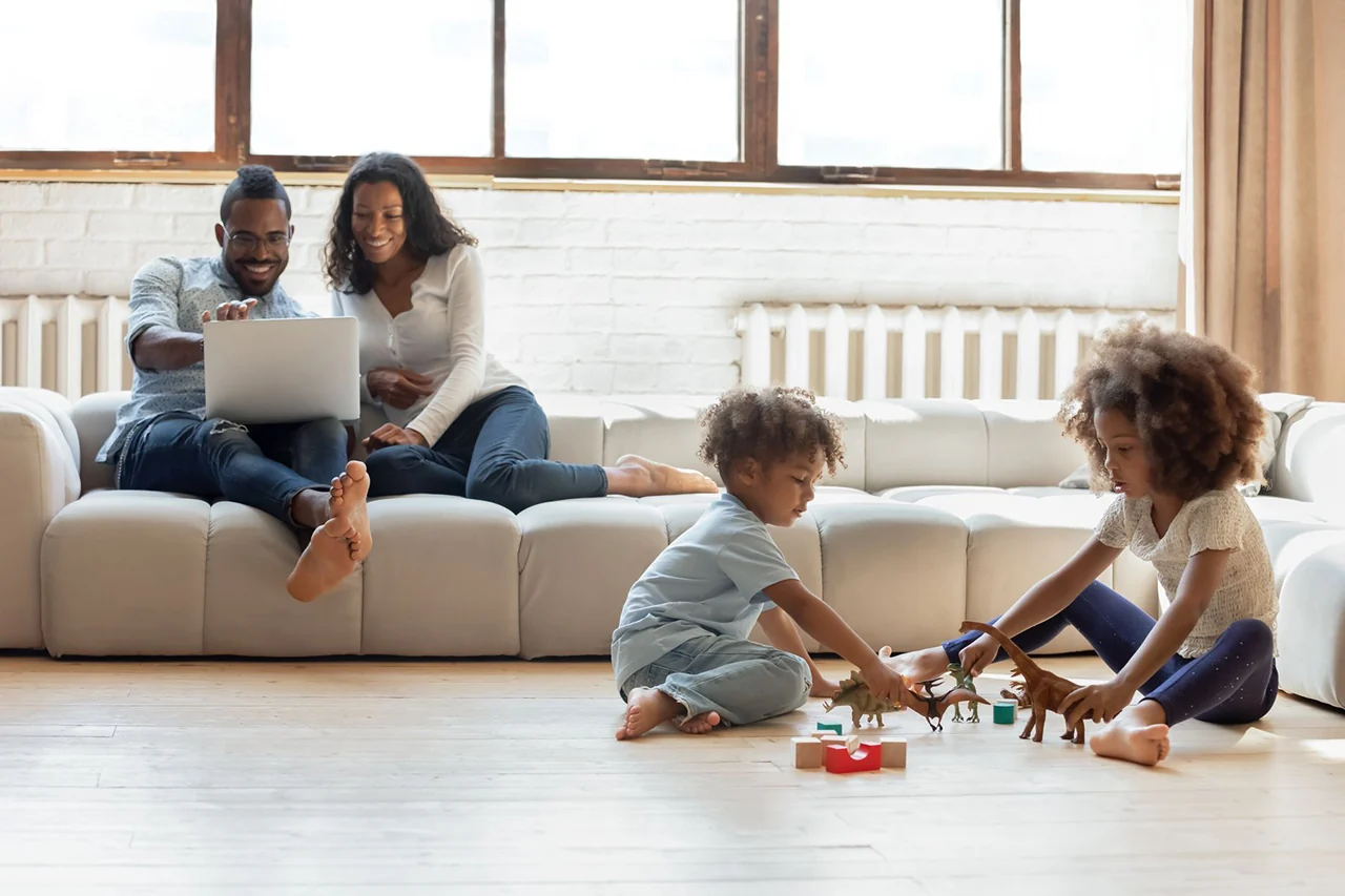 Two parents sit on a sofa with a laptop while two young children play with toy dinosaurs on a wooden floor