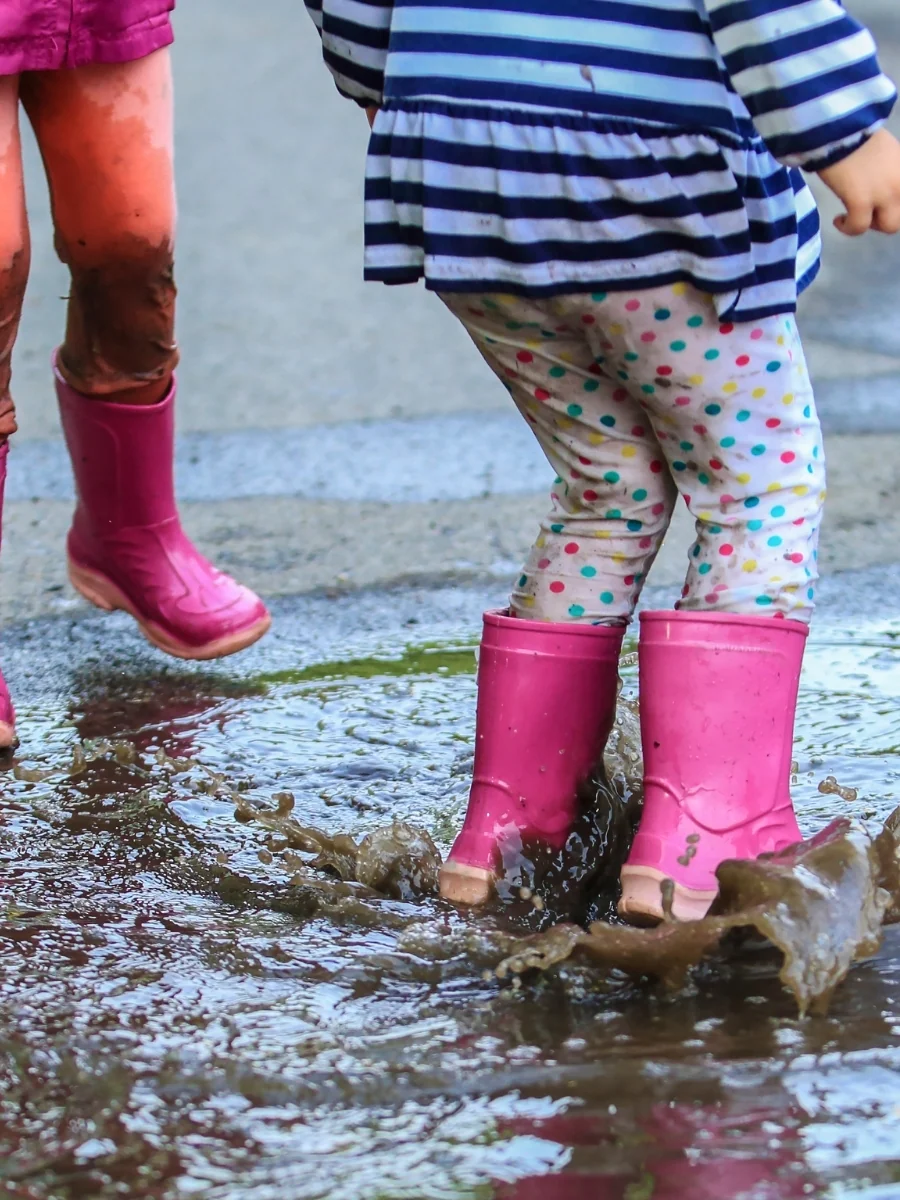 Two children in pink rubber rain boots splashing in a muddy puddle