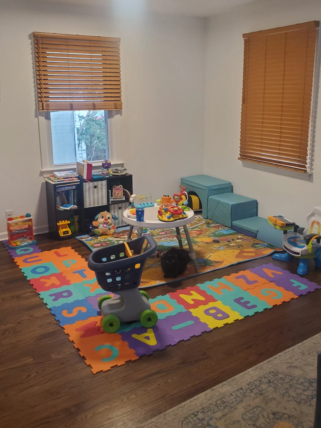 A play area in the corner of a room: a small cube shelf, an activity table and a foam alphabet mat on the floor