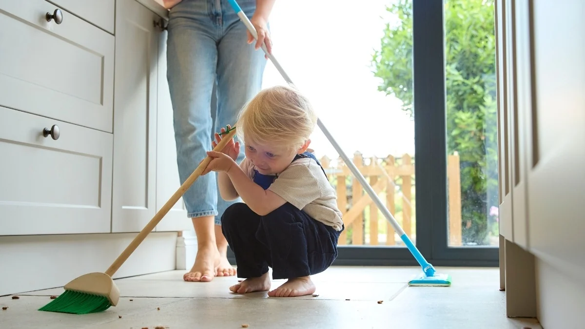 Toddler with a small broom on a kitchen floor while a parent cleans behind them: practical life activities for Montessori