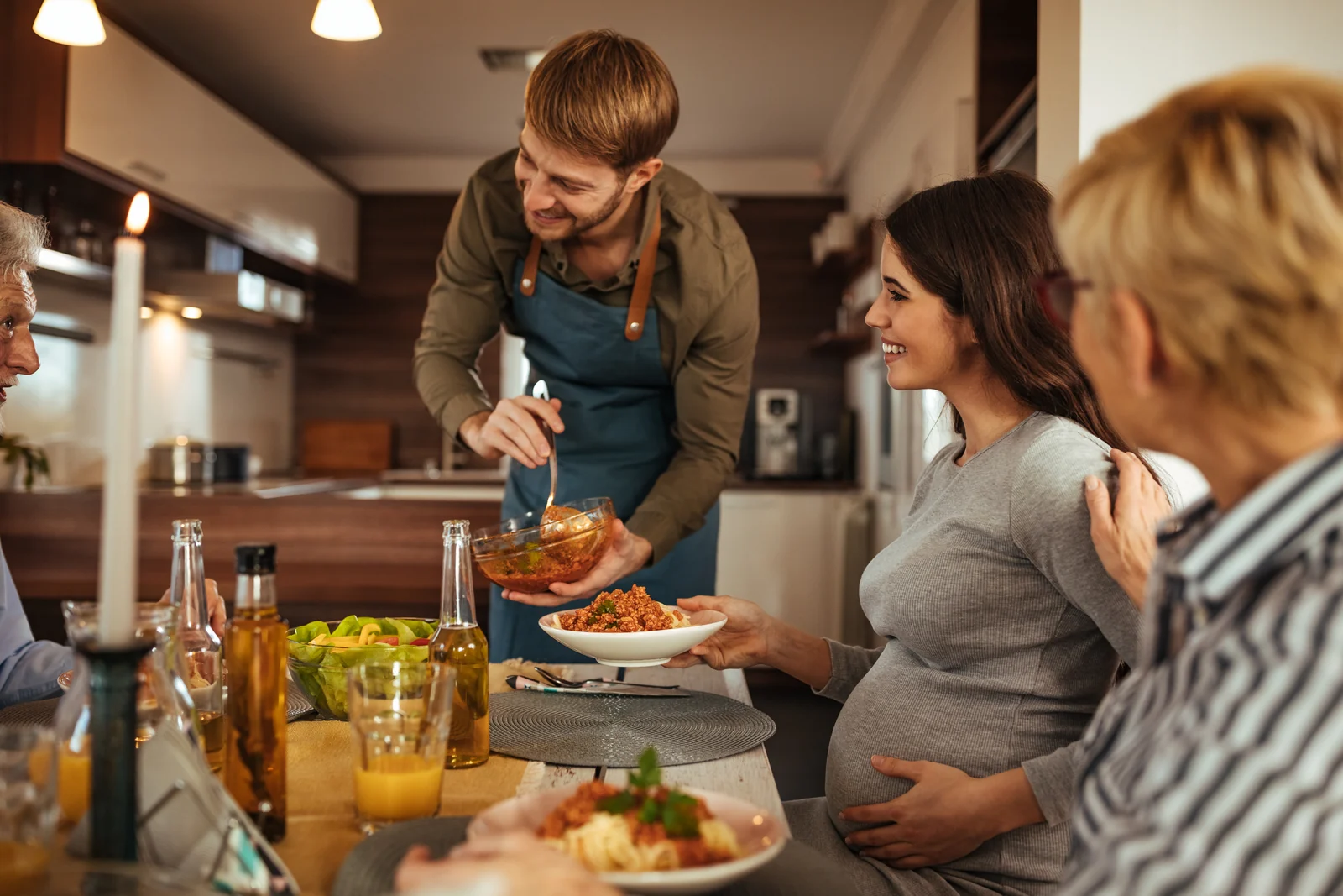 Pregnant person at a family dinner table, listening while a relative serves food