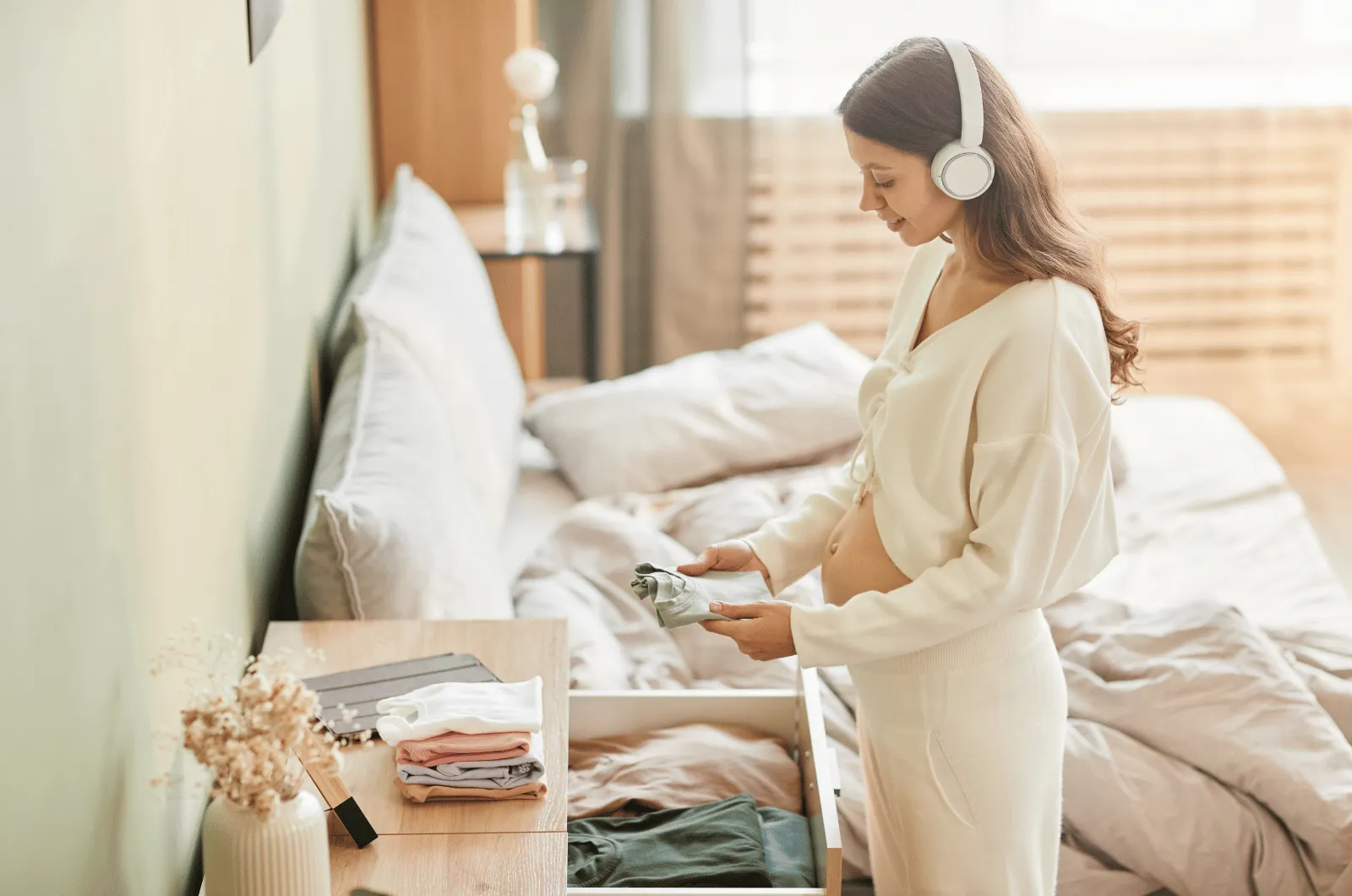 Pregnant parent folding small baby clothes, wearing headphone