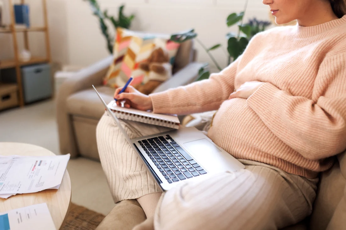 Pregnant woman on the couch with a laptop on her lap, writing notes as she compares baby items