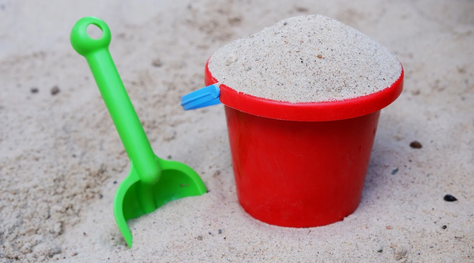 A red toy bucket heaped with sand next to a green plastic shovel standing in pale sand