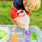 Toddler pouring water from a jug into cups in a clear plastic tub on the grass: sensory activities for toddlers