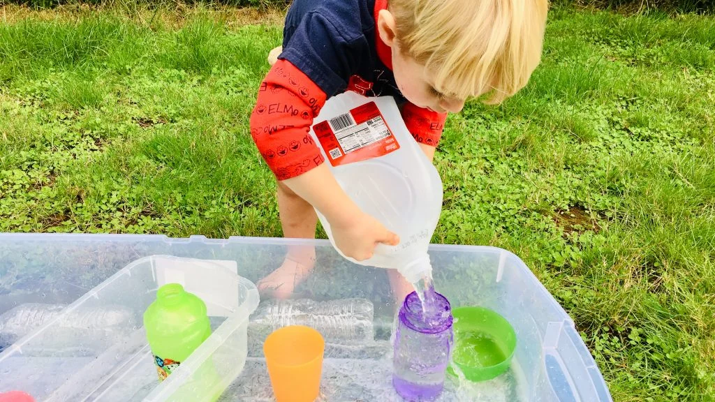 Toddler pouring water from a jug into cups in a clear plastic tub on the grass: sensory activities for toddlers